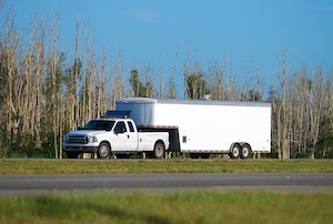 A white medium duty pickup truck towing a long dual axle trailer attached by fifth wheel driving on a NC country highway